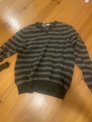 Randig tröja Dressmann i bomull - Snygg randig tröja från Dressmann i storlek S, tillverkad av mjuk pima bomull. Tröjan har mörkgröna och gråa breda ränder, ribbad v-ringad hals och muddar vid ärmslut och nederkant. Perfekt för dig som gillar enkel och stilren design.