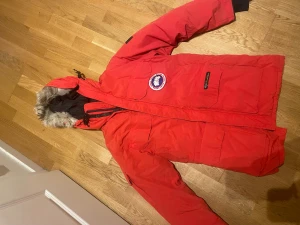 Röd dunparkas från Canada Goose - Säljer en röd Canada Goose parkas med dunfyllning och päls på huvan. Jackan har flera stora fickor, svart innerfoder och klassisk logga på bröstet. Yttermaterialet är polyester och bomull, med nylonfoder. Perfekt för kalla vinterdagar.