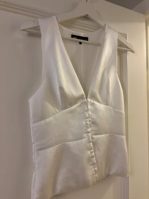 Vit sarin topp från Zara - Snygg vit topp från Zara i satin med djup v-ringning och breda axelband. Toppen har en stilren, figurnära passform och dolda knappar framtill. Perfekt för att lyfta din outfit med en clean och modern vibe. Använd 1 gång, säljer då den ej kommer till användning.