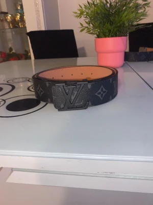 Svart  bälte med monogram - Snyggt svart bälte  med klassiskt monogrammönster och -spänne i mörk metall. Bältet har en bred och stilren design, perfekt för att lyfta din outfit. Materialet är skinn med en lyxig känsla och detaljer i grått.