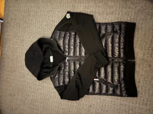 Svart pufferjacka med huva från Moncler - Svart pufferjacka från Moncler med huva och långa ärmar. Jackan har quiltad framsida i glansigt material och släta ärmar samt rygg. Dragkedja framtill och två sidofickor med dragkedja. Moncler-logga på vänster ärm. Perfekt för dig som gillar streetwear.