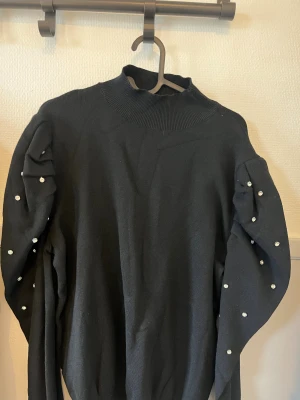 Svart stickad polotröja med pärlor - Snygg svart stickad polotröja med högt ribbat krage och långa ärmar. Ärmarna är dekorerade med pärlor som ger en extra touch. Perfekt för dig som gillar stilrena plagg med lite bling. Tröjan har en loose passform och är superbekväm.