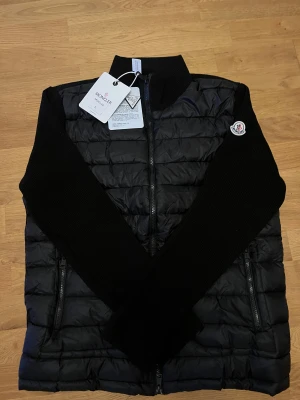 Svart pufferjacka från Moncler - Svart pufferjacka från Moncler med stickade ärmar och quiltad kropp. Jackan har dragkedja framtill, två sidofickor med dragkedja och Moncler-logga på ärmen. Insidan har en cool serietidningsetikett och klassisk Moncler-märkning.