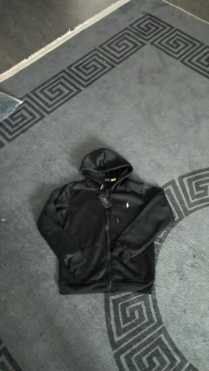 Svart hoodie från Polo Ralph Lauren - Svart hoodie från Polo Ralph Lauren med klassisk vit logga på bröstet. Tröjan har huva med snörning, dragkedja framtill och två fickor. Materialet är mjukt och känns skönt mot huden, perfekt för chill dagar. Snygg och enkel design som funkar året runt.