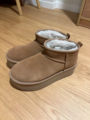 UGG bruna boots med plattform - Säljer ett par bruna UGG boots med plattformssula. Utsidan är i mjuk mocka och insidan är fodrad med fluffig syntetpäls för extra värme. Perfekta för kalla dagar och har en chunky look som är supertrendig just nu.