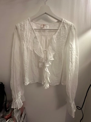 Vit blus med volang från Nelly - Säljer en vit blus från Nelly i storlek 38. Blusen har långa ärmar med knyt vid ärmslut, volangdetaljer längs v-ringningen och framtill, samt dekorativa sömmar upptill. Materialet känns lätt och luftigt, perfekt för en fräsch look.