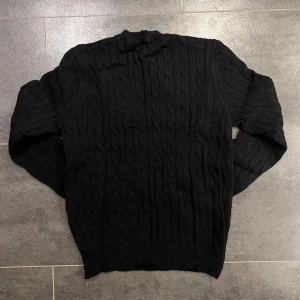 Svart Polo Ralph Lauren - Svart kabelstickad tröja från Polo Ralph Lauren med klassisk rund hals och röd broderad logga på bröstet. Tröjan har långa ärmar och ribbade muddar vid ärmslut och nederkant. Perfekt för dig som gillar stilren och tidlös design.