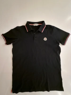 Svart Moncler pikétröja med röd/vit kant - Svart pikétröja från Moncler med klassisk krage och korta ärmar. Tröjan har Moncler-logga på bröstet och snygga rödvita detaljer på krage och ärmslut. Tillverkad i mjuk bomull som känns skön mot huden. Perfekt för en clean och sportig look.