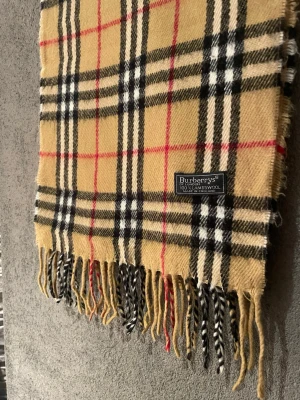 Rutig beige ullväst från Burberrys - Säljer en klassisk rutig väst från Burberrys i beige, svart, vit och röd. Västen är tillverkad i 100% lammull och har fransar längs nederkanten. Den har det ikoniska Burberrys-mönstret och en snygg etikett framtill.