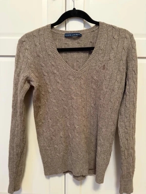 Beige stickad tröja från Ralph Lauren - Snygg beige stickad tröja från Ralph Lauren med klassiskt kabelmönster och v-ringad hals. Tröjan har långa ärmar och ribbade muddar. 80% ull och 20% cashmere.