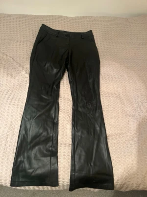 Svarta skinnbyxor från Design by Si - Snygga svarta bootcut-byxor från Design by Si i mjukt skinnmaterial. Byxorna har klassisk midja, bälteshällor och knappstängning framtill. Perfekt för dig som vill ha en cool och stilren look med lite edge. Finns ingen lapp med strl på, men skulle säga att de passar en S-M runt W26/28.