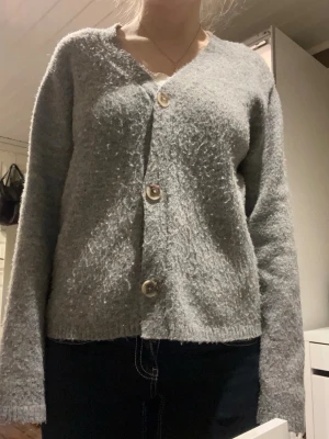 Grå kofta med knappar - Mysig grå kofta med tre stora knappar framtill. Koftan har en lite fluffig och stickad yta, v-ringad hals och långa ärmar. Perfekt för kyliga dagar och enkel att matcha med jeans eller kjol. Lite nopprig.