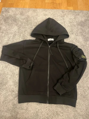 Svart stone island zip up hoodie - Helt ny och oanvänd. Den är storlek M men passar S också. Den har inga fläckar eller någon syn av användning då den är helt oanvänd. Den är varm men samtidigt luftig, 10/10 kofta som jag älskar men jag har XL så den passar inte mig
