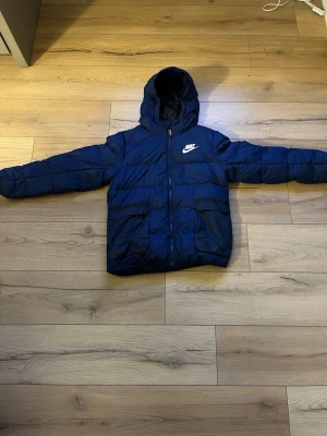 Blå Nike pufferjacka med huva XL - Säljer en blå pufferjacka från Nike i storlek XL. Jackan har huva, dragkedja framtill och två stora fickor. Nike-logga på bröstet och Therma-Fit-teknologi som håller dig varm. Perfekt för kyliga dagar och enkel att matcha med streetwear.
