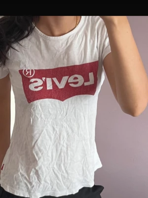 Vit Levi's t-shirt med röd logga - Klassisk vit t-shirt från Levi's med den ikoniska röda loggan tryckt framtill. T-shirten har rund halsringning, korta ärmar och är gjord i mjuk bomull. Perfekt basic-plagg med snygg passform och diskret röd detalj i nederkant.
