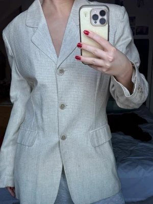 Beige rutig kavaj från JOY - Snygg beige kavaj från JOY med diskret rutigt mönster och klassisk krage. Kavajen har tre knappar framtill, två fickor med lock och är helfodrad. Perfekt för dig som gillar stilrena och tidlösa plagg med en chill vibe.