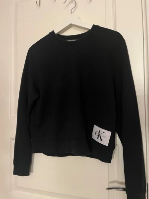 Svart sweatshirt från Calvin Klein Jeans - Svart sweatshirt från Calvin Klein Jeans med rund halsringning och lång ärm. Tröjan har en vit patch med CK-logga nertill på framsidan. Klassisk och enkel design i mjuk bomull, perfekt till chill dagar. Använd 2-3 gånger 