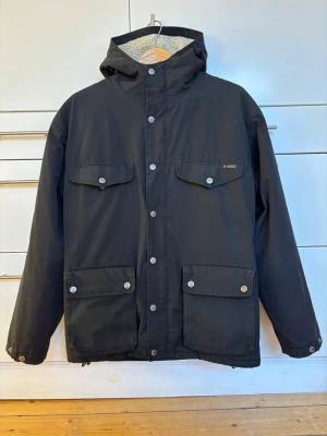 Svart klassisk parkajacka Greenland från Fjällräven storlek XS - Svart klassisk parkajacka Greenland från Fjällräven med huva och fyra stora fickor framtill. Jackan har G-1000-tyg, tryckknappar med logga och ett mjukt teddyfoder. Klassisk Fjällräven-patch på ärmen. Gott skick! Storlek XS