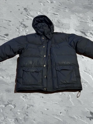 Svart dunjacka från Boomerang XL - Säljer en svart pufferjacka från Boomerang i storlek XL. Jackan har huva, stora fickor framtill och stängs med både dragkedja och knappar. Broderad logga på ärmen och insidan. Perfekt för kalla dagar och riktigt klassisk look.