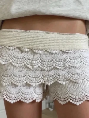 Vita spetsiga shorts med volang - Säljer ett par vita shorts med bred resår i midjan och flera lager dekorativ spets med volangkant. De har en feminin och bohemisk vibe, perfekta för varma dagar. Materialet är bomull med spetsdetaljer och de är högmidjade för en snygg siluett.