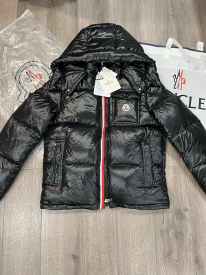 Moncler jacka  - Moncler montbeliard med glansig finish, huva och ikonisk logga på bröstet. Jackan har en rödvit randig dragkedja framtill och praktiska fickor med dragkedja. Perfekt för dig som vill ha en snygg och varm jacka med streetkänsla.
