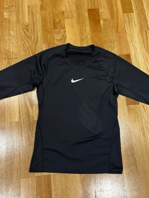 Svart Nike långärmad träningströja - Svart långärmad träningströja (compression shirt)  från Nike med klassisk vit swoosh-logga på bröstet. Tröjan är tillverkad i ett syntetmaterial som andas och har en rund halsringning. Perfekt för dig som gillar sport och vill ha en stilren look.