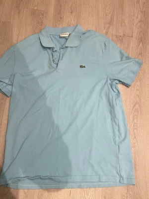 Ljusblå piké från Lacoste - Säljer en ljusblå pikétröja från Lacoste med klassisk krage och korta ärmar. Tröjan har knappar framtill och den ikoniska broderade krokodilloggan på bröstet. Perfekt för dig som gillar stilrena och sportiga plagg.