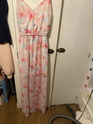 Blommig långklänning från Gina Tricot - Säljer en superfin långklänning från Gina Tricot i storlek 34. Klänningen har ett ljust blommigt mönster i rosa, blått och gult, v-ringad omlott upptill och tunna axelband. Materialet är mjukt och lätt polyester, perfekt för varma dagar.