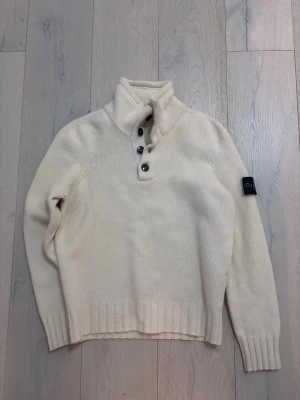 Vit stickad polotröja från Stone Island - Säljer en vit stickad polotröja från Stone Island med hög krage och fyra svarta knappar framtill. Tröjan har långärmad design och ribbade muddar vid ärmslut och nederkant. Klassisk Stone Island-logga på vänster ärm. Perfekt för dig som gillar stilrena och exklusiva plagg.