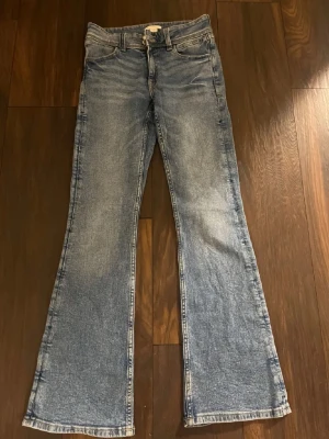 Blå bootcut jeans från Gina Tricot - Säljer ett par blå bootcut jeans från Gina Tricot i storlek 164. Jeansen har klassisk femficksdesign, ljus tvätt och snygga broderade detaljer på bakfickorna. Perfekt för dig som gillar retrovibbar och vill ha ett par jeans med lite extra sväng i benen.