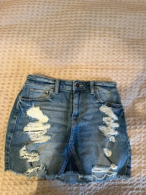 Blå jeanskjol från Hollister (W26) - Säljer en blå high-rise jeanskjol från Hollister med vintage stretch. Kjolen har slitna detaljer, fransig nederkant och klassiska fickor både fram och bak. Perfekt för dig som gillar en avslappnad och trendig look.