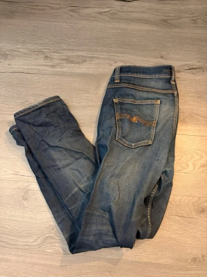 Blå jeans från Nudie Jeans - Säljer ett par klassiska blå jeans från Nudie Jeans med snygga slitningar och kontrastsömmar. Modellen har fem fickor och orange broderi på bakfickorna. Jeansen är raka i benen och har en normal passform. Materialet är robust denim i bomull.