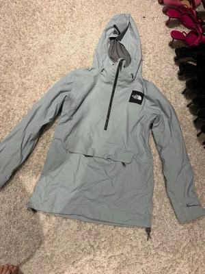 Ljusblå skidjacka från The North Face - Säljer en ljusgrå vindjacka från The North Face med huva och half zip framtill. Jackan har en stor ficka på magen och justerbar dragsko i huvan. Perfekt för blåsiga dagar och outdoor-äventyr. Enkel och stilren design med logga på bröstet.