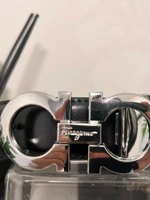 Ferragamo bälte - Säljer ett stilrent svart bälte från Ferragamo med en ikonisk, blank silverfärgad spänne. Bältet är tillverkat i slätt skinn och har Ferragamo-loggan ingraverad på spännet. Perfekt accessoar för att lyfta din outfit med en lyxig touch. 110cm