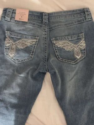 Blå bootcut jeans med paljettdetaljer - Snygga ljusblå jeans från Gina Tricot i bootcut-modell med låg midja. Fikorna är broderade med silvriga paljetter i ett coolt mönster. Jeansen har slitningar framtill och är helt nya. Perfekt för dig som gillar detaljer!