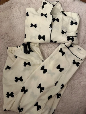 Vit pyjamasskjorta + byxor  med svarta rosetter - Säljer en vit pyjamasskjorta med svarta rosetter och svarta kantdetaljer. Skjortan har klassisk krage, knappstängning och en ficka framtill. Materialet är mjukt och har en lätt struktur. Matchande byxa med rosett i midjan ingår.