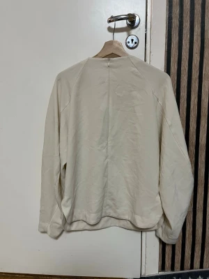 Vit/Beige långärmad tröja i bomull - Snygg vit:beige långärmad tröja med rund hals och enkel design. Tillverkad med en dragkedja i rugen som kan öppnas halvvägs. Perfekt för dig som gillar stilrena och neutrala plagg. Passar dig som vill ha en basic tröja med avslappnad passform.