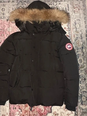 Svart Canada Goose dunjacka med päls - Svart dunjacka från Canada Goose med ikonisk patch på ärmen och fluffig päls runt huvan. Jackan har dragkedja och knappar framtill, två stora fickor och quiltad design. Insidan är fodrad med blankt tyg och jackan är perfekt för kalla vinterdagar.