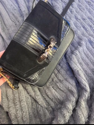 Ny pris 700 - Snygg svart axelväska med guldfärgad kedja och flera fack. Väskan har en mix av mocka och skinn, samt detaljer i blankt material och ett coolt mönstrat spänne framtill. Perfekt storlek för det viktigaste och stilren design som sticker ut.