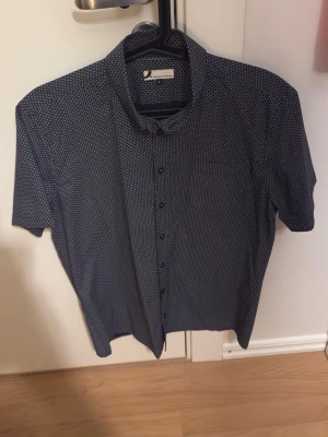 Mönstrad blå skjorta från Dressmann - Säljer en blå skjorta från Dressmann med vitt geometriskt mönster. Skjortan har korta ärmar, klassisk krage och knäppning framtill. Perfekt för dig som gillar stilrena men ändå unika plagg. Materialet känns lätt och skönt mot huden.