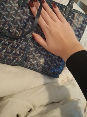 Blå axelväska med mönster - Säljer en snygg blå axelväska  med det klassiska grafiska mönstret i vitt och brunt. Väskan har stilrena detaljer och rejäla handtag, perfekt för dig som gillar exklusiva accessoarer. Materialet är skinn och väskan har vita sömmar som kontrast.