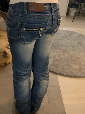 Coola jeans  - Säljer ett par blå jeans från D-stag med coola slitningar och en unik dragkedja på bakfickan. Jeansen har normal passform och klassisk femficksdesign. Materialet är robust denim och färgen är snyggt tvättad med ljusare partier framtill och baktill.