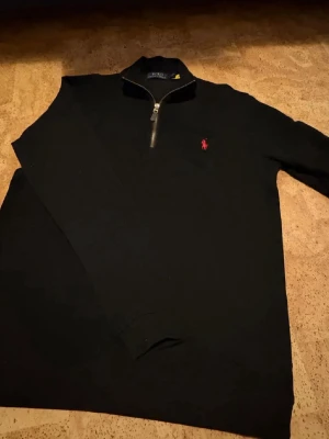 Svart half zip tröja från Polo Ralph Lauren - Svart långärmad tröja från Polo Ralph Lauren med half zip och röd broderad logga på bröstet. Tröjan har en klassisk krage och är gjord i mjuk bomull, perfekt för en chill och stilren look.