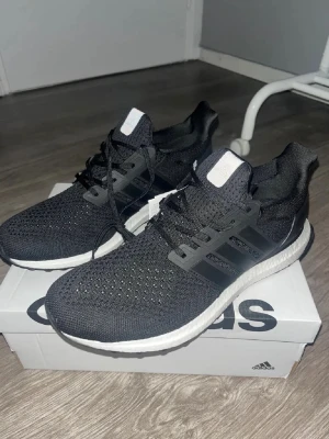 Adidas Ultraboost svarta sneakers - Svarta Adidas Ultraboost sneakers med stickad ovandel och klassiska tre ränder på sidan. Skorna har snörning och en vit, responsiv Boost-sula som ger grym komfort. Perfekta för dig som gillar stilrena och sportiga skor med modern känsla.
