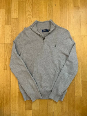 Halv dragkedja Ralph lauren tröja - Snygg grå stickad tröja från Polo Ralph Lauren med halv dragkedja och klassisk krage. Tröjan har långärmad design och diskret logga broderad på bröstet. Perfekt för lager-på-lager och chill stil under höst och vinter.