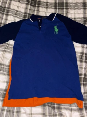 Blå och orange pikétröja Polo Ralph Lauren - Snygg pikétröja från Polo Ralph Lauren i blått och orange med grönt broderat logomärke på bröstet och stor POLO-text på ryggen. Tröjan har korta ärmar, klassisk krage med knappar och vita detaljer vid kragen. Perfekt för dig som gillar sportig och färgstark stil.