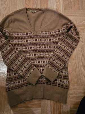 Beige mönstrad ulltröja Barbour - Säljer en beige stickad ulltröja från Barbour med v-ringning och långärmad design. Tröjan har ett snyggt mönster i svart, rosa och grönt över bröst och ärmar samt ribbade muddar. Perfekt för dig som gillar klassisk stil med en twist.