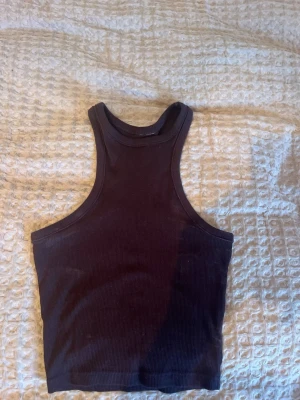 Mörkbrun ribbad linnetopp - Säljer en mörkbrun ribbad linnetopp med hög hals och racerback-design. Toppen är ärmlös och har en tight passform som framhäver axlarna. Perfekt för att styla med jeans eller kjol under varma dagar. Materialet är mjukt och stretchigt för extra komfort. Ingen lapp kvar, men passar en S.