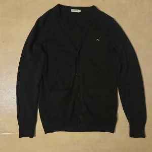 Svart kofta från J.Lindeberg, storlek S/M - Svart kofta från J.Lindeberg i storlek S/M. Tillverkad i 100% bomull, med v-ringad hals, knappar framtill och två fickor. Diskret J.Lindeberg-logga broderad på bröstet. Perfekt att slänga på över en t-shirt för en clean look.