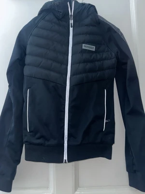Svart pufferjacka från McKenzie XS - Säljer en svart pufferjacka från McKenzie i storlek XS. Jackan har huva, vita detaljer vid dragkedja och fickor samt quiltad framsida. Tillverkad i polyester och har långa ärmar med diskreta reflexdetaljer på axeln. Perfekt för dig som vill ha en sportig och clean look.